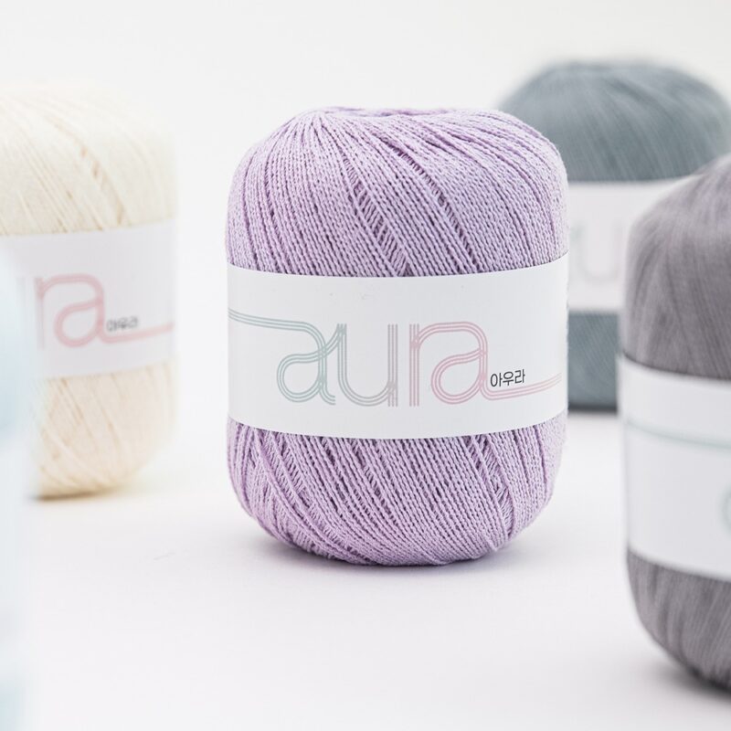 Lavender Aura yarn skein in focus on a white surface, with beige and gray skeins blurred in the background.