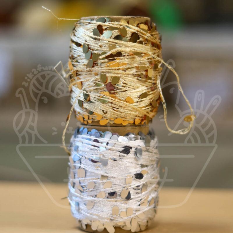 Two decorated cardboard rolls stacked vertically, wrapped in twine with gold and silver sequin confetti, forming a craft ornament with a loop of string protruding.